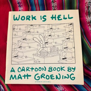 May include: A cartoon book titled "WORK IS HELL" by Matt Groening. The cover features a black and white cartoon of a rabbit-like character in a chaotic office setting. The book is on a colorful patterned textile.