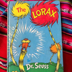 May include: A vintage book cover featuring a bright red and yellow illustration of the Lorax, a fictional character created by Dr. Seuss. The cover is blue with a white background and features the text "The Lorax" in yellow letters.