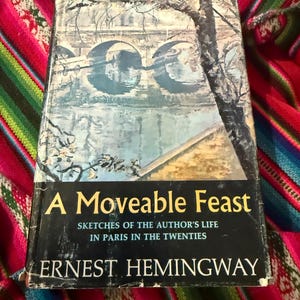May include: A well-worn copy of "A Moveable Feast" by Ernest Hemingway. The book cover features a watercolor painting of a bridge over a river. The title is in gold lettering, with the author's name below.