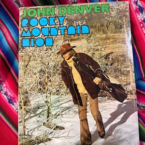 May include: Album cover for John Denver's "Rocky Mountain High" featuring a photo of the artist in a snowy landscape. The album cover has a green and blue title with a photo of John Denver wearing a brown hat and coat.