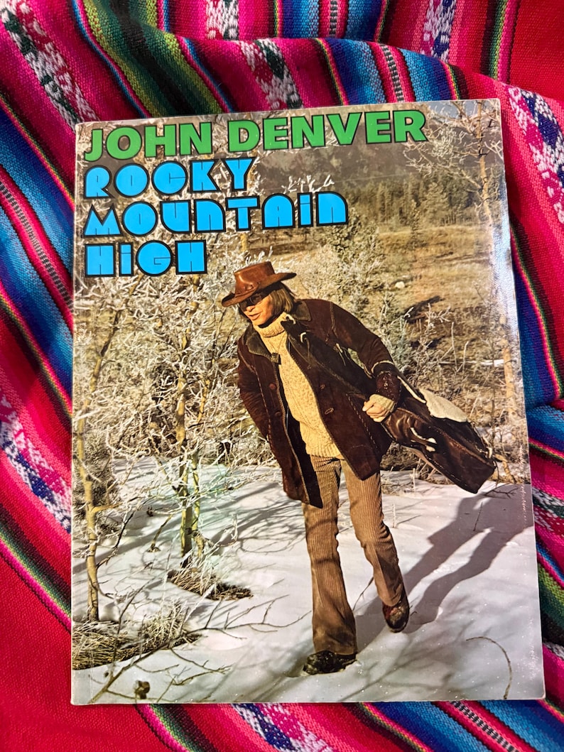 May include: Album cover for John Denver's "Rocky Mountain High" featuring a photo of the artist in a snowy landscape. The album cover has a green and blue title with a photo of John Denver wearing a brown hat and coat.