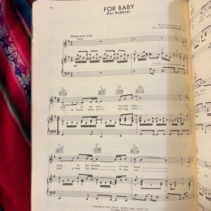 May include: Open sheet music titled "FOR BABY (For Bobbie)" with musical notes and lyrics. The page is from a vintage songbook, with text including "Words and Music by H. J. DEUTSCHENDORF, JR." and copyright information. The background is a colorful textile.