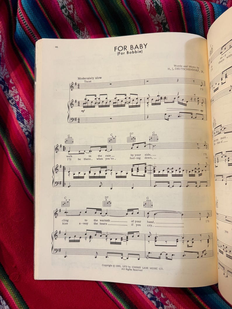 May include: Open sheet music titled "FOR BABY (For Bobbie)" with musical notes and lyrics. The page is from a vintage songbook, with text including "Words and Music by H. J. DEUTSCHENDORF, JR." and copyright information. The background is a colorful textile.