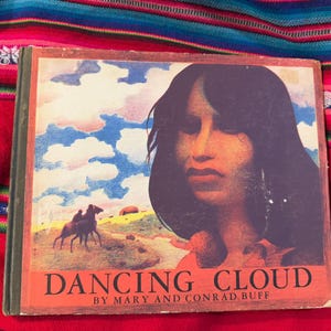 May include: Vintage book titled "Dancing Cloud" by Mary and Conrad Buff. The cover features a portrait of a woman with long dark hair, a landscape with a horse, and a blue sky with clouds. The book is set against a colorful textile.