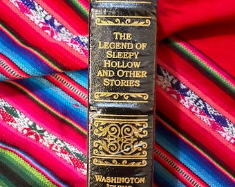 May include: A black book titled "The Legend of Sleepy Hollow and Other Stories" by Washington Irving. The book's spine features gold decorative accents and text. The book is wrapped in clear plastic and rests on a colorful textile.