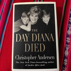 May include: A white book with a black cover and the title "The Day Diana Died" by Christopher Andersen. The book cover features a black and white photo of Diana, Princess of Wales, with her two sons, Prince William and Prince Harry.