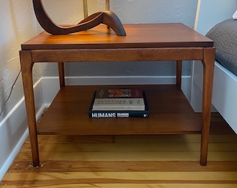 May include: A mid-century modern style wooden side table with a lower shelf. The table is a warm brown color and has a rectangular top. A lamp with a curved wooden base and a beige lampshade sits on top. Two books are on the lower shelf.