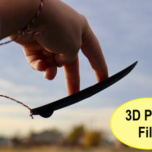 May include: A black 3D printed object is held by a finger, with a string attached to it. The object is shaped like a thin, flat blade. The background is a blurry image of a blue sky and green grass. The text "3D Print File" is in a yellow oval.
