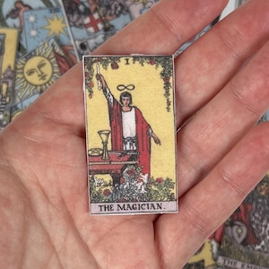 May include: A rectangular tarot card featuring "The Magician" with a red robe, a table with a chalice, and the infinity symbol above the figure's head. The card has a yellow background and is surrounded by other tarot cards.