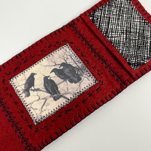 May include: A red felt bookmark with a black stitched border and a printed image of three crows perched on a tree branch. The bookmark has a black and white patterned pocket.