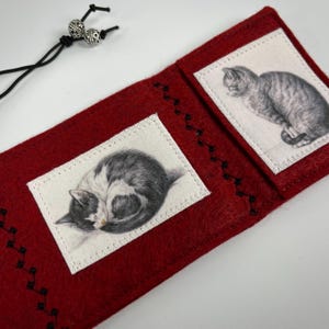 May include: A red felt bookmark with two rectangular patches featuring detailed pencil drawings of cats. The bookmark has black stitching and a black cord with silver beads.