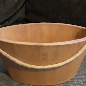 May include: A wooden bucket with a rope handle. The bucket is oval shaped and has a natural wood finish.