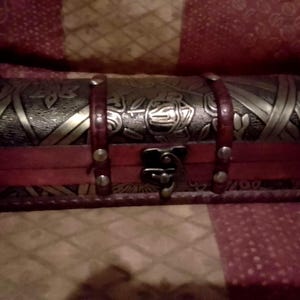 Op de afbeelding: Een decoratieve houten kist met een donkerbruin gewelfd deksel en een rode basis. Het deksel is versierd met goudkleurige bloemen- en geweven ontwerpen, vastgezet met rode leren riemen en een metalen sluiting. Opbergdoos in vintage stijl.