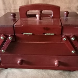 May include: A wooden sewing box with a hinged lid, a drawer, and two side compartments. The box is painted a deep red color and has a handle on the top.