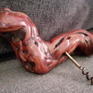 May include: A wooden corkscrew with a red-brown finish. The corkscrew is shaped like a twisted branch and has a metal worm.