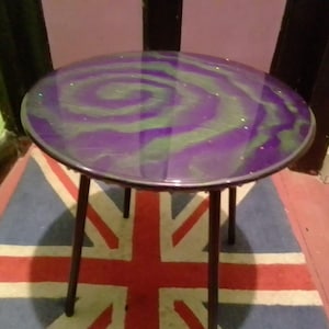 Puede incluir: Una mesa auxiliar redonda con una tapa de resina epoxi de color púrpura y verde con un remolino y patas de metal negro. La mesa está sobre una alfombra con un patrón de Union Jack.
