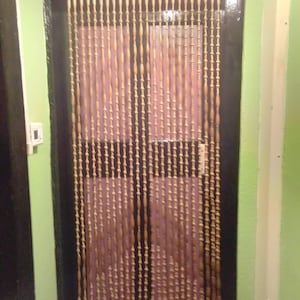 Könnte beinhalten: Ein Perlenvorhang aus hellbraunen Holzperlen. Der Vorhang hängt in einem Türrahmen mit dunklem Rahmen vor einem hellrosa Hintergrund. Der Türrahmen ist dunkelbraun und die Wand hellgrün.
