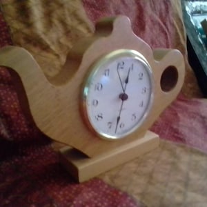 Puede incluir: Un reloj de madera con forma de tetera con una esfera blanca y ribete dorado. El reloj tiene manecillas negras y un cuerpo de madera marrón claro. El reloj está sobre una base rectangular.