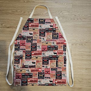 May include: A red, white, and black BBQ-themed apron with a repeating pattern of BBQ-related graphics and text. The apron has a white neck strap and waist ties. The design includes images of cows, BBQ signs, and the words "BBQ" and "Barbeque Heaven."