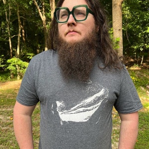 Könnte beinhalten: Ein dunkelgraues T-Shirt mit einem weißen Grafikdesign. Die Person auf dem Bild trägt eine große, quadratische Brille mit grünem Rahmen. Der Hintergrund ist eine natürliche Umgebung im Freien mit Bäumen und Grün.