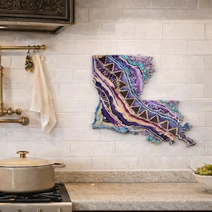 May include: A colorful, abstract wall art piece in the shape of Louisiana. The artwork features shades of purple, blue, and gold, with a textured, layered design. It is mounted on a white brick wall in a kitchen setting.