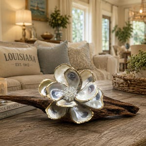 May include: A decorative flower sculpture crafted from iridescent oyster shells with gold-painted edges and a central cluster of small white pearls, mounted on a piece of dark, weathered driftwood, creating a natural and organic aesthetic.