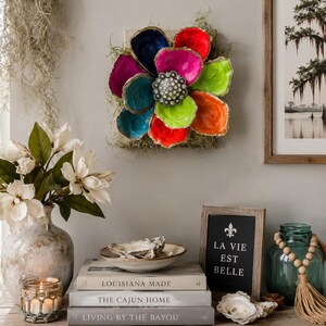 May include: A wall-mounted floral art piece crafted from painted oyster shells in vibrant shades of blue, red, magenta, lime green, and orange, arranged around a central silver-toned metallic embellishment with clear faceted stones.