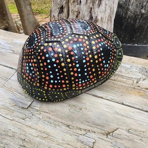 May include: A painted turtle shell with a black base and a pattern of colorful dots in orange, yellow, and blue. The shell is resting on a weathered wooden surface, showcasing the intricate design and texture.