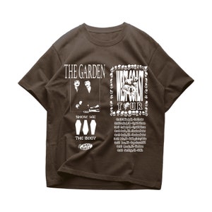 May include: A brown t-shirt with a white graphic print of a band called "The Garden" with the text "West Coast Tour" and a list of tour dates. The graphic also includes the text "Show Me The Body" and a logo for "Deli Records".