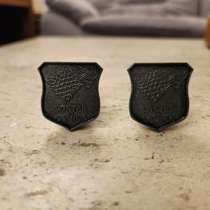 May include: A pair of black cufflinks featuring a stylized direwolf head with the words "Winter is Coming" on each cufflink.