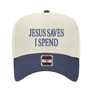May include: A baseball cap with a cream-colored crown and a navy blue brim. The text "JESUS SAVES I SPEND" is embroidered in navy blue across the front. The cap has a curved brim and a button on top.