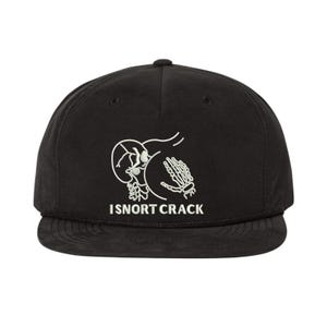 May include: Black baseball cap with a flat peak. The cap displays a white graphic of a skull, a buttock, and a skeleton hand. The text "ISNORT CRACK" is printed below the graphic in white.