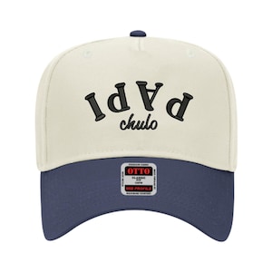 May include: A baseball cap with a cream-colored crown and a navy blue brim. The cap features the words "PAPI chulo" embroidered in black. The cap has a curved brim and a button on top.