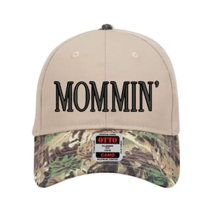 May include: A beige baseball cap with the word "MOMMIN'" embroidered in black. The brim features a camouflage pattern. The cap has a classic fit and is made of premium fabric.