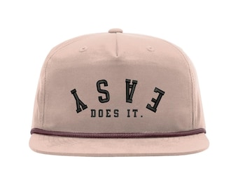 May include: A light pink flat-brimmed cap with the words "EASY DOES IT" embroidered in black. The cap has a brown rope detail along the brim.