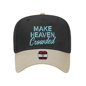 Puede incluir: Una gorra de béisbol negra con visera color canela que presenta el texto bordado en azul claro "MAKE HEAVEN Crowded" en la parte delantera. Una pegatina rectangular con el logotipo "OTTO" en rojo y negro está centrada en la visera.