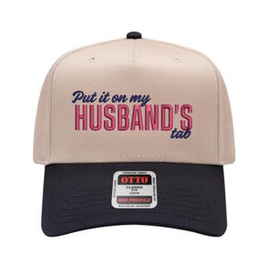 Puede incluir: Gorra de béisbol beige y negra con el texto "Put it on my HUSBAND'S tab" en rosa y azul. La gorra tiene una visera curva y un botón en la parte superior. La marca "OTTO" es visible.