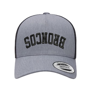 May include: A gray and black trucker hat with the word "BRONCOS" embroidered in black on the front. The hat has a black mesh back and a gray front panel. The hat has a "The Authentic Snapback" label on the back.