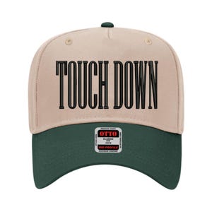Op de afbeelding: Een beige en donkergroene baseballcap met de woorden "TOUCH DOWN" in vette zwarte letters. De cap heeft een gebogen klep en een knoop aan de bovenkant. Het merk "OTTO" is zichtbaar.