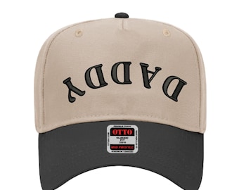 May include: A beige and black baseball cap with the word "DADDY" embroidered in black. The cap has a curved brim and a small tag on the front.
