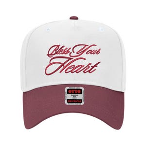 May include: A white baseball cap with a maroon brim and top button. The phrase "Bless Your Heart" is embroidered in red script across the front. The cap has a mid-profile fit and a tag that reads "OTTO".