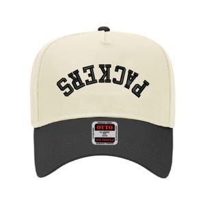 May include: A cream-colored baseball cap with a black curved brim. The word "PACKERS" is arched across the front in black block letters. A small rectangular patch with text is on the lower front.