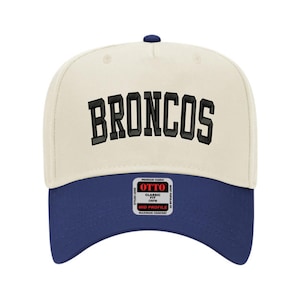 May include: A baseball cap with a cream-colored crown and a navy blue brim. The word "BRONCOS" is embroidered in black block letters across the front. The cap has a curved brim and a small navy blue button on top.