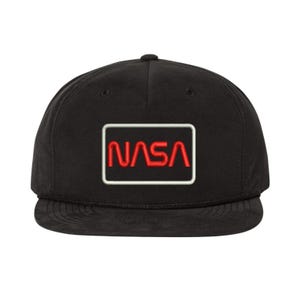 May include: Black baseball cap with a flat brim. The cap features a rectangular patch with the NASA logo in red letters inside a white border. The hat has a structured crown and a curved brim.