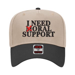 May include: A beige and black baseball cap with the text "I NEED MORAL SUPPORT" embroidered on the front. The word "MORAL" is crossed out with a red X.
