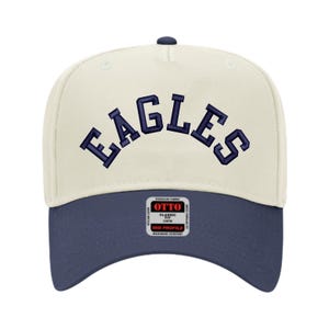 May include: A cream and navy blue baseball cap with the word "EAGLES" arched across the front in navy blue. The cap has a curved brim and a small tag on the front.