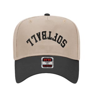 May include: A beige and black baseball cap with the word "SOFTBALL" embroidered in black across the front. The cap has a curved brim and a small black button on top. The brand label "OTTO" is visible on the front.