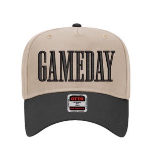 May include: A beige and black baseball cap with the word "GAMEDAY" embroidered in large, black, block letters across the front. The cap has a curved brim and a small tag on the lower front.