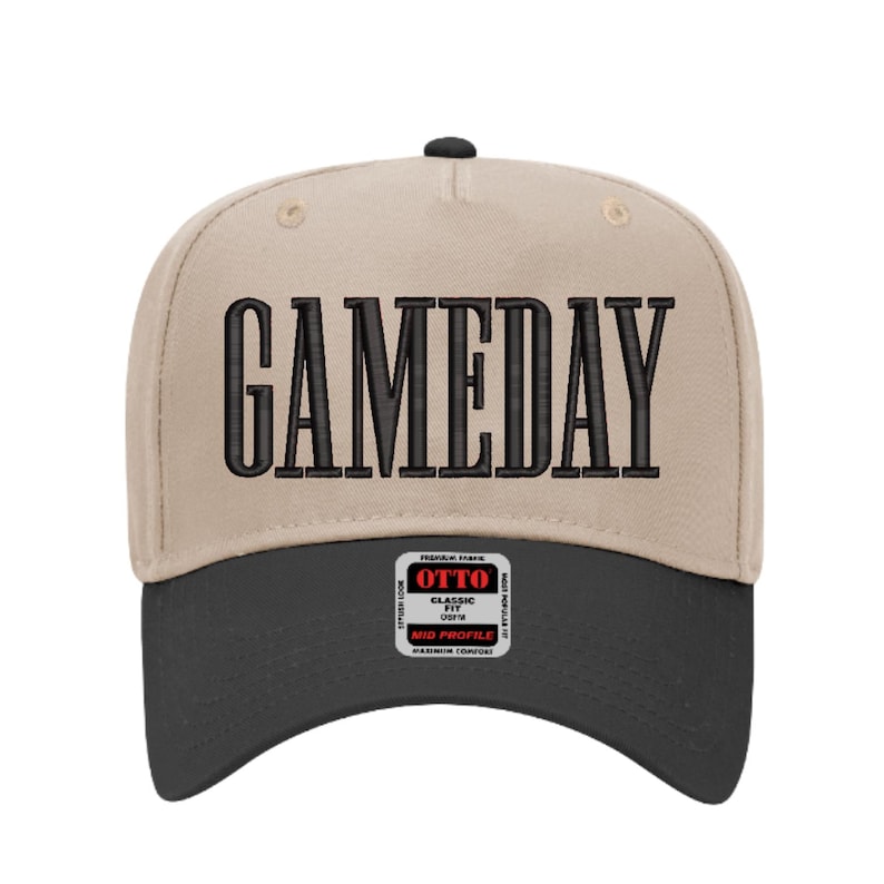 May include: A beige and black baseball cap with the word "GAMEDAY" embroidered in large, black, block letters across the front. The cap has a curved brim and a small tag on the lower front.
