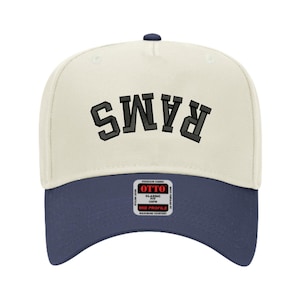 May include: A baseball cap with a cream-coloured crown and a navy blue brim. The word "RAMS" is embroidered in a dark grey, block-style font across the front. The cap has a curved brim and a small navy blue button on top.